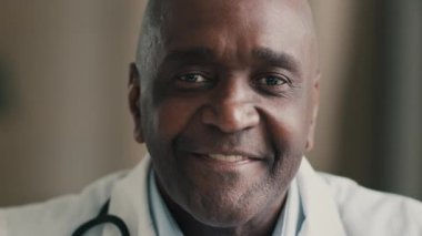 Portre yetişkin gülümseyen Afrikalı Amerikalı yaşlı erkek terapist göz doktoru doktor doktor üniformalı dişçi hastane kliniğinde erkek pratisyen kardiyolog sağlık konseptinde oturuyor.