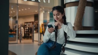 Ortadoğulu bayan iş kadını Arap kökenli kız öğrenci telefonda alışveriş yapıyor alışveriş merkezinde oturuyor mobil uygulamalar kullanıyor internetten dijital haber videoları okuyor.