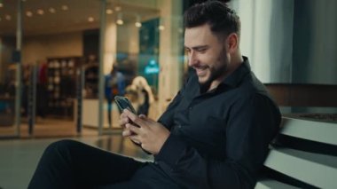 Siyah takım elbiseli beyaz bir adam başarılı bir işadamı alışveriş merkezinde cep telefonu uygulamaları ile oturuyor. İnternetten kıyafet alıyor, internet indirimleri kazanıyor, uzaktan iletişim kuruyor.