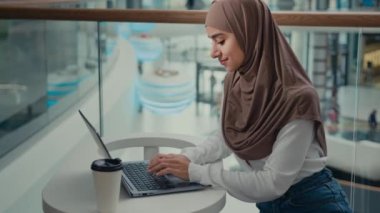 Müslüman kız Arap kız öğrenci alışveriş merkezinde oturuyor. Laptop kullanıyor. İnternette çalışıyor. İslamcı kadın tüketici sosyal ağlarda iletişim kuruyor.