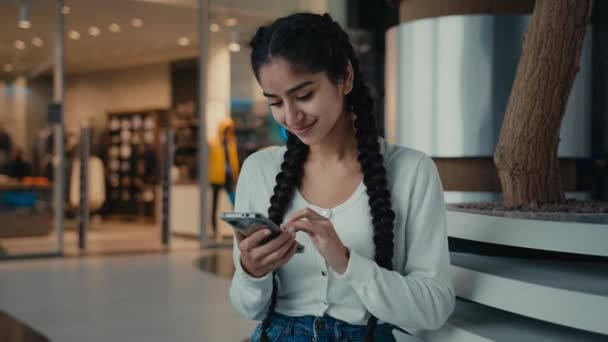 魅力的なアラビア語の若い女性の学生中東の女の子は ショッピングモール見て座っている画面のテキストメッセージビジネスチャットに遠くの通信のための携帯電話を使用してインターネットストアで注文を行う — ストック動画