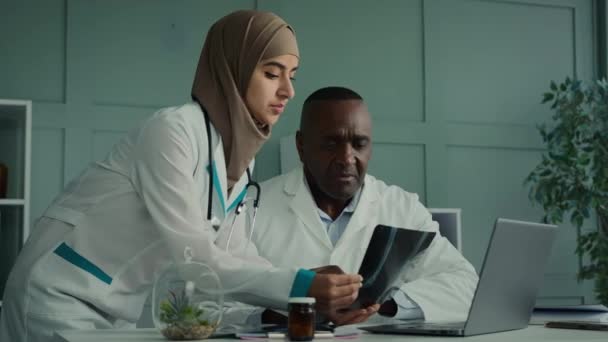 2人の医療関係者は アフリカの高齢男性医師に骨画像とMriスキャンを示すRoentgen若いアラビア人女性放射線医を見て放射線診断X線結果の専門知識 — ストック動画