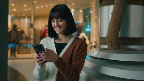 笑顔20代の女性女性女性が屋内ショッピングモール内の携帯電話の画面を見て座っているスマートフォンの笑顔でネットアプリでブラウジングスクロールソーシャルメディアでチャット電子商取引オンラインで購入 — ストック動画