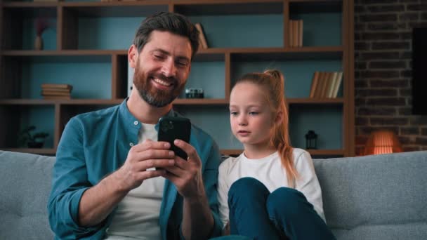 かわいい小さな赤ちゃんの女の子子供は思いやりのある若い父親の男と居心地の良いソファに休んでいるスマートフォンでゲームをプレイメディアコンテンツを見ます携帯アプリを使用して携帯電話で買い物をオンラインで一緒に家族の自撮り — ストック動画