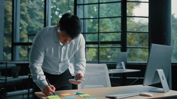 高级亚洲人企业家商人站在现代办公室的桌旁用手机在便条上写任务组织文书工作计划多任务工作集思广益 — 图库视频影像