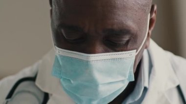 Tıbbi maskeli Amerikalı Afro-Afrikalı doktor solunum cihazını kovid-19 virüsünden koruyor tecrit edilmiş erkek doktorda oturuyor reçeteli ilaç test sonuçları yazıyor.