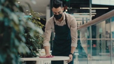 Kafe çalışanı, erkek garson, kafeterya müdürü, tıbbi maskeli ve önlüklü küçük işletme sahibi restoran açılmadan önce tozlu kirli masayı silip, covid 19 koronavirüs salgını temizledi.