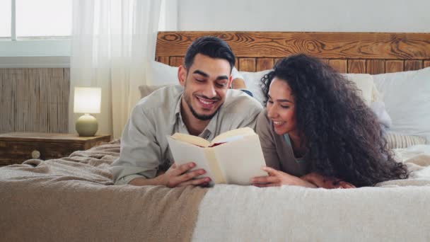 多种族的多种族夫妇女人和留胡子的男人躺在床上看书 新婚夫妇在家里看文艺小说 未来的父母会在课本中为孩子取一个名字 — 图库视频影像