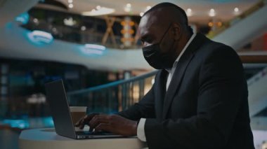 Orta yaşlı yetişkin adam yönetici patron şirket CEO iş adamı iş adamı kafeteryada dizüstü bilgisayarla çalışıyor tıbbi yüz maskesi takıyor COVID-19 coronavirus salgını