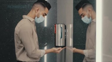 Havaalanındaki kamu şirketinde tıbbi maske takan etnik erkek işadamı WC otomatik alkol dezenfektanı sabun jeli otomobil antibakteriyel makinesi ile ellerini yıkıyor. Covid-19 önle