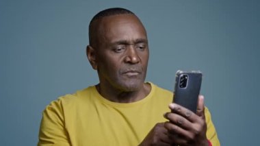 Olgun adam gri arka planda stüdyoda duruyor cep telefonuyla sosyal ağlara takılıyor spor atletleri internetteki fiziksel egzersizleri izliyor akıllı telefon ekranına işaret ediyor kafa sallıyor
