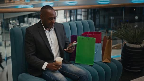 アフリカの成人男性中年ビジネスマンとショッピングバッグモール店でコーヒーティーカップブラウジングスクロール電話アプリオンライン購入注文スマートフォン割引で予約する — ストック動画