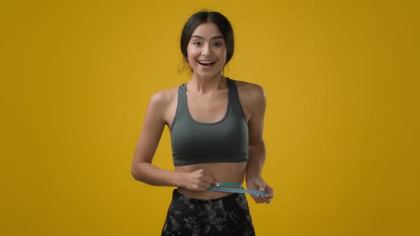 インドの強いですスリムスポーツ女の子運動選手女性とともにメジャーテープ測定腰サイズでスタジオ黄色の背景幸せな喜びで成功した達成良い健康体形状緩い体重トレーニング — ストック動画
