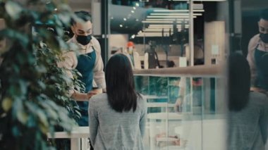 Erkek garson kafeterya müdürü önlüklü ve tıbbi maskeli adam bayan ziyaretçi müşteriyle konuşuyor. Masadaki kıza kahve, çay ve kapuçino getir.