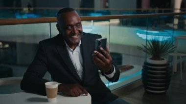 Orta yaşlı erkek girişimci, kafede görüntülü görüşme yapıyor. Kahve içen Afro-Amerikalı iş adamı telefon konferansı ile konuşuyor.
