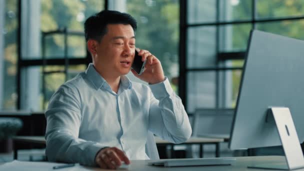 自信を持って大人40代のビジネスマンアジアの日本人男性コンサルタントは オフィスの職場に座っている携帯電話の無線を話す5G接続遠くの携帯電話の呼び出しは リモートでビジネスコンサルタントの職業を交渉 — ストック動画