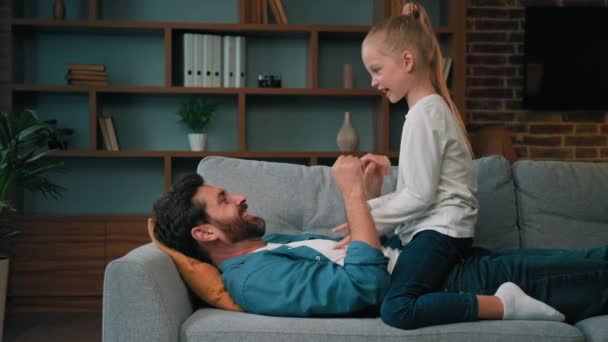 可爱的小女孩和爸爸在舒适的沙发上玩耍 一起玩乐 感觉很好 有爱心的高加索男人爸爸拥抱女儿快乐童年的概念 — 图库视频影像