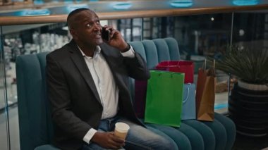 Orta yaşlı Afrikalı bir iş adamı alışveriş poşetleriyle alışveriş merkezindeki dükkanda telefonla konuşuyor. Satın almalar hakkında konuşuyor. Akıllı telefonla Kara Cuma indirimi hakkında konuşuyor.