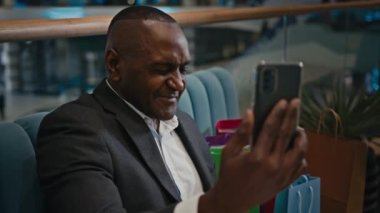 Alışveriş merkezindeki alışveriş torbaları ile orta yaşlı Afro-Amerikalı bir erkek işadamı video konferans konferansında uzaktan satış konuşmaları yapıyor.