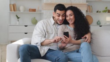 Latin Hintli, çok ırklı, çok ırklı bir çift. Evli bir adam ve kadın, telefon takibi, akıllı telefon uygulaması kullanarak kanepede sarılıyorlar. İyi haberler okuyorlar.