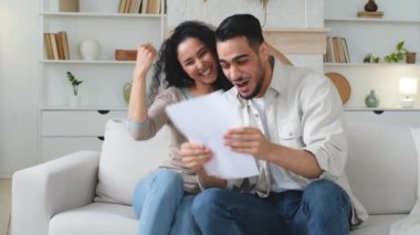 Coşkulu, etnik çeşitlilikte bir çift kanepeye oturur ve mutlu bir şekilde evrak kâğıdı okurlar. Harika bir galibiyetle birlikte. Banka kredisi onayıyla kazançlı bir iş fırsatı sunarlar.