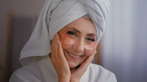 Positive portrait happy senior lady with anti wrinkle skin hydrogel collagen patches under eyes smiling to camera old 60s Caucasian woman with towel on head enjoy morning beauty rituals with skincare
