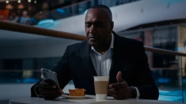 Ethnic African American middle-aged businessman in cafe table mature man CEO with mobile phone business chatting relaxing eat cake eating pie drink tea drinking coffee break with smartphone browsing