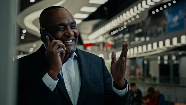Smiling African American middle-aged man talking mobile phone in company shopping mall. Cheerful adult mature businessman enjoy cellphone call happy entrepreneur calling smartphone indoors speak laugh
