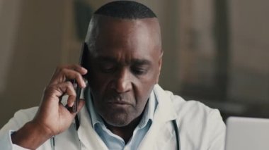 Afrika kökenli Amerikalı doktor tıp uzmanı cep telefonuna danışmanlık yapıyor yaşlı terapist genel pratisyen hekimler sağlık hizmetlerini online olarak tartışıyor klinikteki modern bilgisayarı kullanıyor