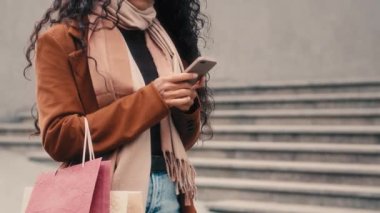 Tanınmayan kadın alışverişçisi, modaya uygun ceketli, şık eşarplı, elinde alışveriş çantası paketleri taksicilik uygulamaları ve akıllı telefon hizmetleri olan bir kız.