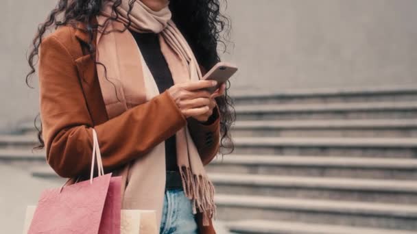 不认识的女购物者不认识的女孩穿着时髦的外套时尚的围巾站在城市里拿着购物袋包用智能手机购买出租汽车应用程序服务 — 图库视频影像