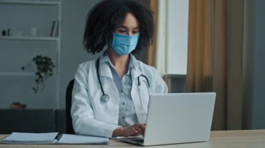 Üniformalı Afrikalı kadın doktor hastane kabinesinde dizüstü bilgisayarda çalışıyor uzaktan elektronik muayene hastaları sanal tıp uygulamasında test sonuçları alıyor.