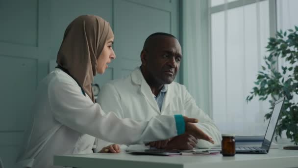医療関係者2人の専門医の同僚アラビアの若い女性セラピストは アフリカのシニア男性と電子健康テストの結果を議論オンラインデジタル診断を行うコンピュータアプリケーションを使用します — ストック動画