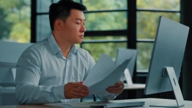 Yorgun Asyalı işçi müdürü ofiste oturup rutin evrak işleriyle uğraşıyor sözleşme belgelerini okuyor bütçe faturalarını internetten analiz ediyor iş planı belgelerini inceliyor profesyonel kağıt denetimi