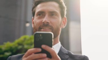 İş portresi genç mutlu iş adamı dışarıda güneş ışığında duruyor telefonla e-posta okuyor mobil uygulama kullanıyor online mağazada sipariş veriyor sosyal ağda geziniyor akıllı telefondan uzaktan sohbet ediyor.