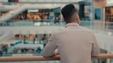 Düşünceli, düşünceli, girişimci, iş adamı, resmi gömlekli, tıbbi yüzlü, koruyucu maskeli, alışveriş merkezindeki ofis maskesinde bekleyen insanlara bakıyor.