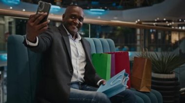 Orta yaşlı erkek Afrikalı erkek alıcı müşteri müşteri blogcu alışveriş merkezindeki video görüşmesinde oturan alışveriş merkezindeki video blogcu etkileyicisi akıllı telefondan fotoğraf satın alarak fotoğraf çekiyor.