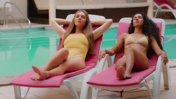 魅力的な若い多民族ヒスパニック系白人女性の友人観光客モデル水着でリラックス一緒に日光浴プールスパホテル近くのサンラウンジャーで夏の豪華な休暇を楽しむ — ストック動画