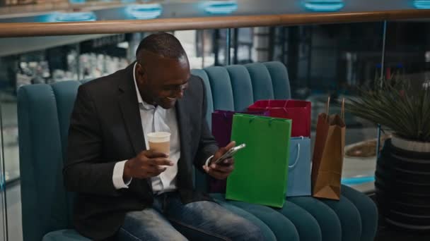 非洲中年男人顾客顾客在沙发上的购物中心购物时 提着购物袋笑着看有趣的视频和手机视频聊天喝茶 — 图库视频影像