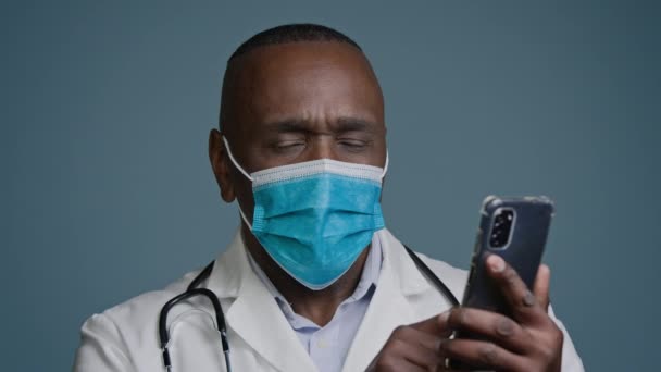 保護マスクに閉じ込められた高価な思慮深いアフリカ系アメリカ人男性医師は インターネットポンダーに関する新しい情報は リモートで患者をオンラインで相談医療モバイルアプリケーションを使用電話研究を保持します — ストック動画