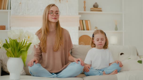 蓮の花の位置でソファに家に座っている小さな子供の女の子と白人女性のお母さんは一緒に瞑想目を閉じた 家族瞑想母親は瞑想を教えて小さな娘可愛いです赤ちゃん子供の息 — ストック動画