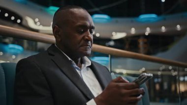 Afrikalı iş adamı cep telefonlu akıllı telefonlara göz atıyor orta yaşlı bir adam cep telefonu kullanıyor cep telefonu teknolojisi kullanıyor içeride bedava WI-FI bağlantısı kuruyor koltukta oturmuş internet telefonlarına bakıyor.