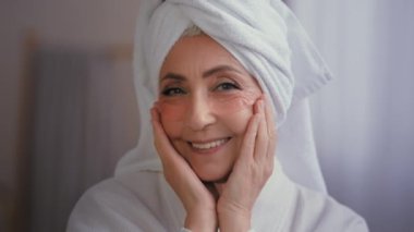 Kapalı portre mutlu yaşlı bayan kırışıklık önleyici cilt hidrojel kolajen bantları kamera altında gülümsüyor. Kafasında havlu olan 60 'lı yaşlardaki beyaz kadın sabah güzellik ayinlerinde cilt bakımı yapıyor.
