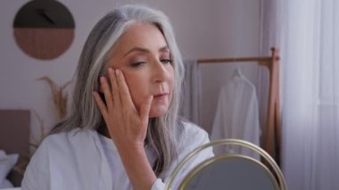 50 'li yaşlarda, gri saçlı, cilt neminden zevk alan yaşlı bir kadın aynadaki yansıma yüz kremine bakıyor. 60' lı yaşlardaki olgun kadınlar kozmetik sonuçlarına göre yüz estetiği yanaklarına dokunuyor.
