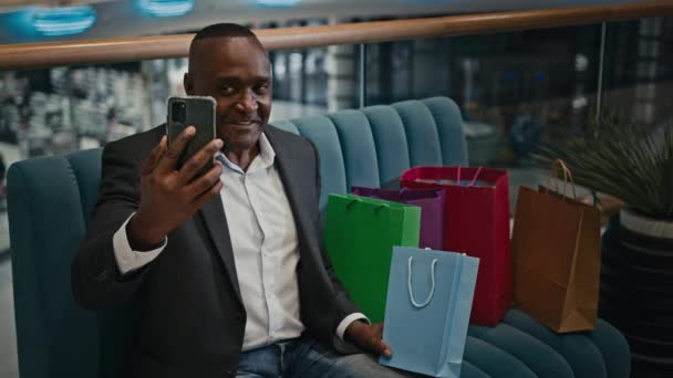 中年笑顔アフリカの成人男性と電話でチャットショッピングモールでの電話ショッピングバッグの購入を示すモバイル会議チャットストアでの話男性消費者バッグの購入について話す — ストック動画