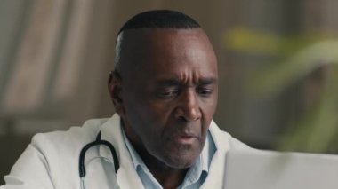 Portre çalışanı olgun 50 'li yaşlarda Afro-Amerikan doktor hastane ofisinde modern bilgisayar danışmanlığı yaparak modern sağlık hizmetleri teknolojisindeki hastalara mesaj yazıyor.