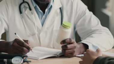 Bilinmeyen etnik Afrikalı erkek doktor doktor doktor pratisyen hekim klinik masasında hasta hastalara danışmanlık yapıyor ilaç şişesini ilaç haplarıyla birlikte tutuyor vitaminler reçeteli tıbbi sevk formu yazıyor.