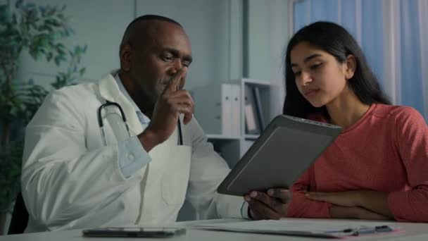 アフリカ系アメリカ人男性医療従事者が病気の女性患者に話すデジタルタブレットで健康診断結果を示す推薦疾患治療を与えるアラビア人女性はクリニックで経験豊富な大人の医師に耳を傾ける — ストック動画