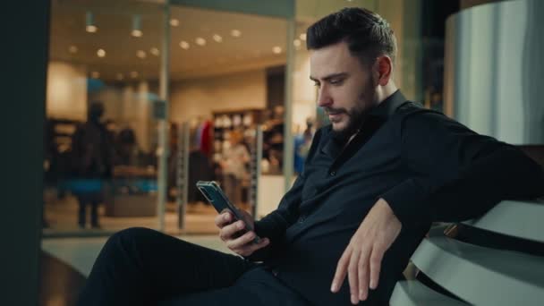 ビジネスマンCaucasian男ひげを生やした男はショッピングビジネスセンターに座っている携帯電話でSmsメッセージを読むソーシャルネットワークでオンラインチャットスマートフォンアプリを使用して注文するモバイルウェブサイトを閲覧 — ストック動画