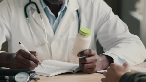 不知名的非洲裔医生医生全科医生坐在办公室诊疗所的桌子前咨询病人 拿着药丸瓶维生素写笔记开处方 — 图库视频影像